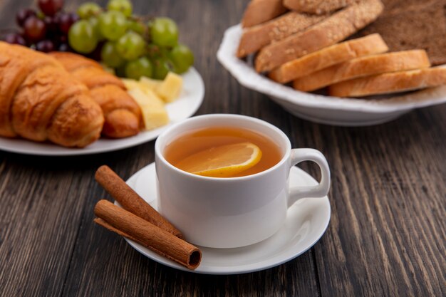 Side view of cup of hot toddy with cinnamon on saucer and croissants with grapes and cheese slices and breads in plates on wooden background