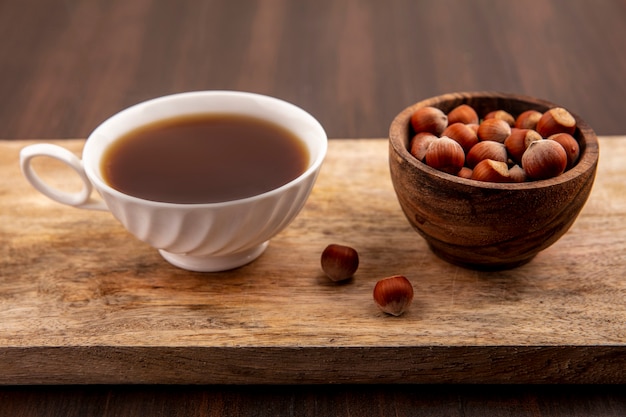 Side view of cup of tea and bowl of nuts on cutting board on wooden background