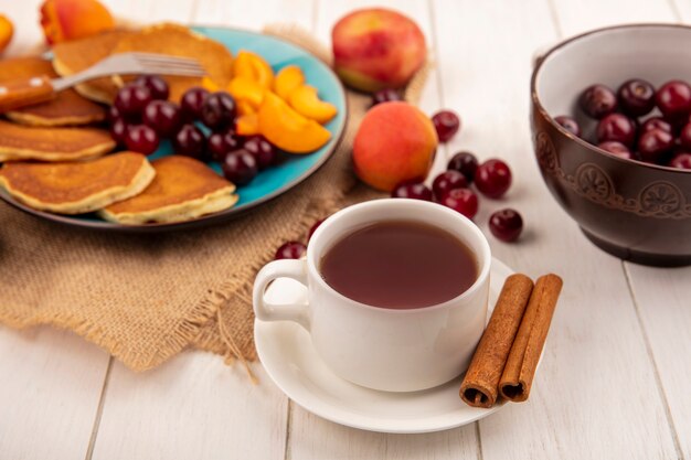 Side view of cup of tea and cinnamon on saucer and pancakes with cherries and apricot pieces in plate and apricots cherries pear on sackcloth and bowl of cherries on wooden background