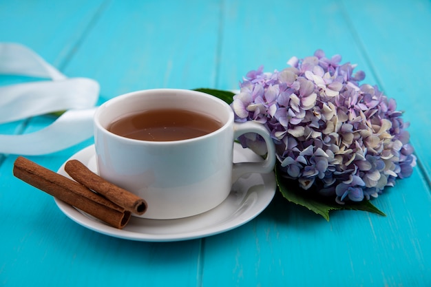 Side view of cup of tea and cinnamon on saucer with flower and ribbon on blue background