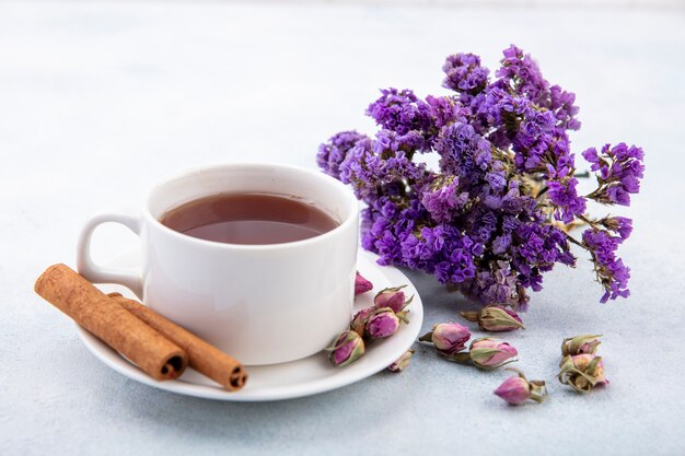 Side view of cup of tea and cinnamon with flowers on saucer and on white surface