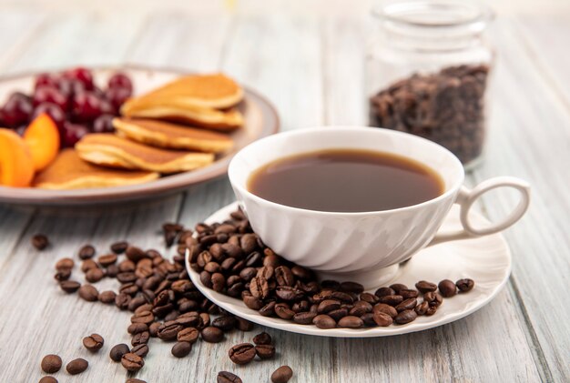 Side view of cup of tea and coffee beans on saucer with plate of pancakes and cherries and apricot slices with jar of coffee beans on wooden background