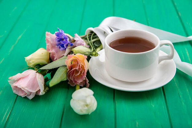 Side view of cup of tea on saucer and flower bouquet on green background