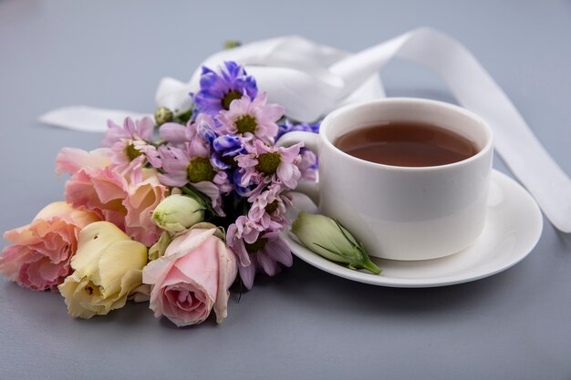 Вид сбоку чашки чая на блюдце и цветы с лентой на сером фоне