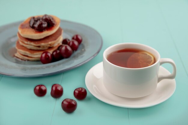 Side view of cup of tea on saucer and pancakes with cherries in plate and on blue background