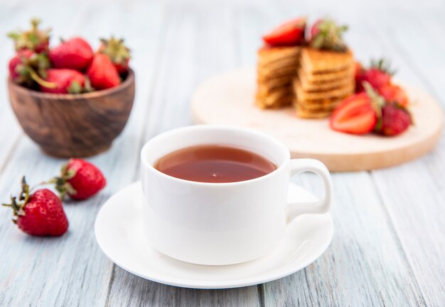 Side view of cup of tea on saucer and waffle biscuits with strawberries in plate and bowl on wooden surface