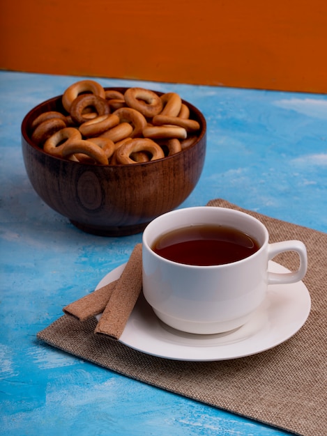 Side view of a cup of tea served with a bowl of bread rings on blue