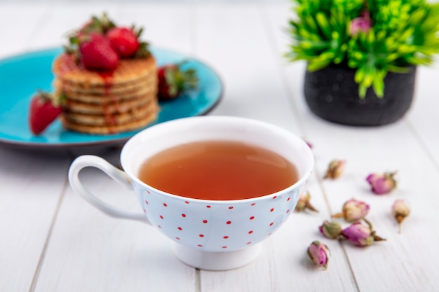 Side view of cup of tea and waffle biscuits with strawberries in plate and flowers on wooden surface