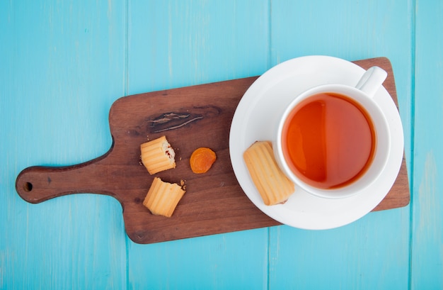 Side view of a cup of tea with biscuits and dried apricot on wood cutting board on blue