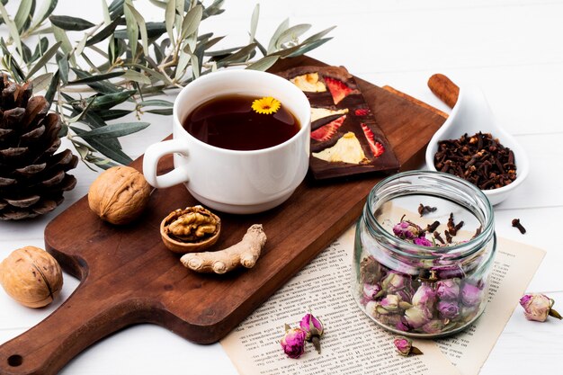 Side view of a cup of tea with chocolate bar with dried fruits and whole walnuts on wood cutting board, dry rose buds in a glass jar and clove spice on white