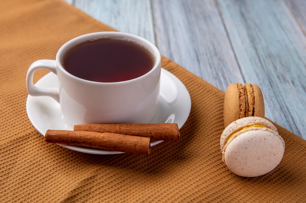 Side view of cup of tea with cinnamon and macarons on a brown towel on a gray surface