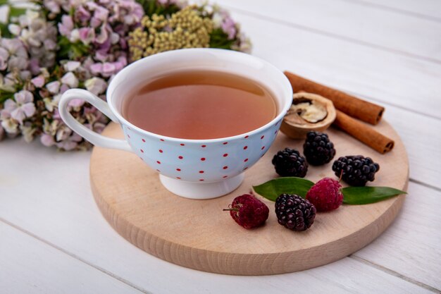 Side view of cup of tea with cinnamon nuts raspberries and blackberries on a cutting board with flowers on a white surface