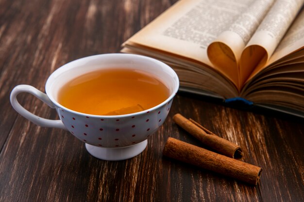 Side view of cup of tea with cinnamon and an opened book on a wooden surface