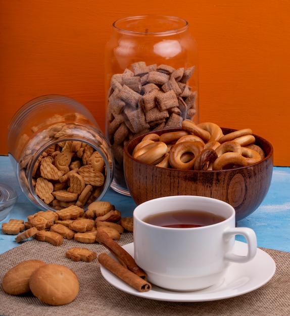 Side view of a cup of tea with cinnamon sticks glass jar of cookies and a bowl with russian bagels on the table