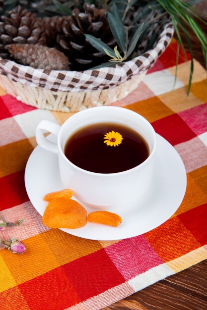 Side view of a cup of tea with dried apricots and pine cones in a basket on plaid tablecloth