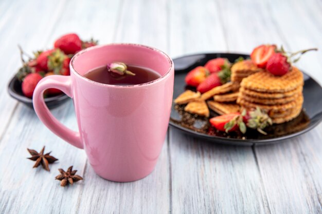 Side view of cup of tea with flower in it and waffle biscuits with strawberries in plates on wooden surface