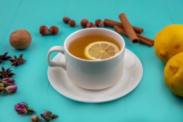 Side view of cup of tea with lemon slice and cinnamon with nuts walnut lemons and flowers on blue background