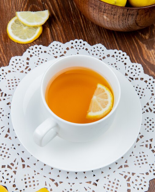 Side view of cup of tea with lemon slice in it on paper doily and lemons on wooden background