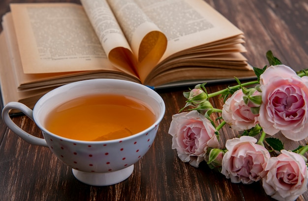 Side view of cup of tea with open book and pink roses on wooden surface