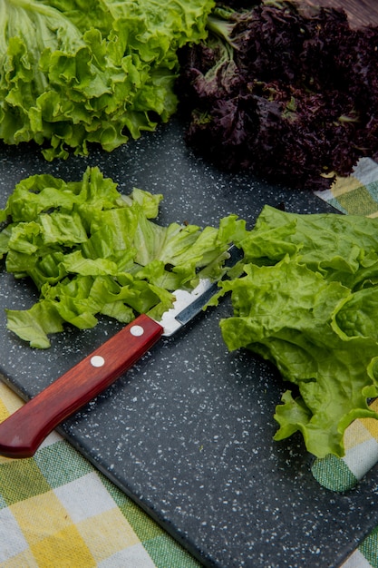 Vista laterale di lattuga tagliata con coltello sul tagliere e uno intero con basilico sul panno plaid