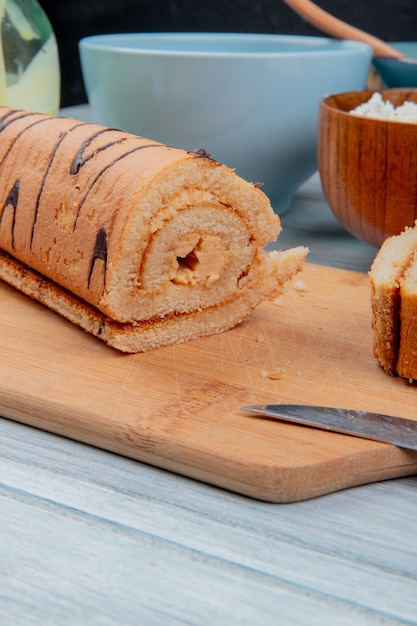 Free photo side view of cut roll with knife on cutting board with condensed milk cottage cheese cereals on wooden surface