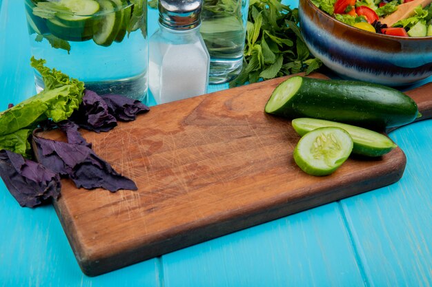 Side view of cut and sliced cucumber on cutting board with basil lettuce mint vegetable salad detox water and salt on blue table