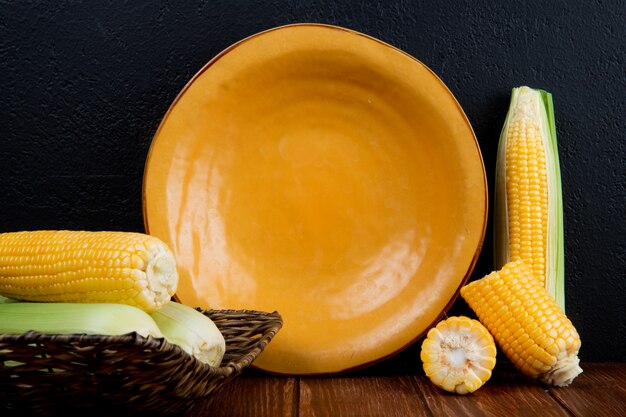 Side view of cut and whole corn cobs and empty plate on wooden surface and black surface