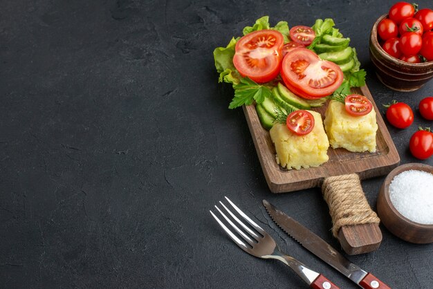 Side view of cut whole fresh tomatoes and cucumbers cheese on wooden board cutlery set salt on the left side on black surface