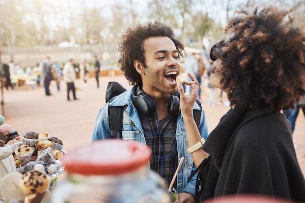 Side-view of cute african-american couple in love having fun in park during food festival, standing near counter and picking something to eat.