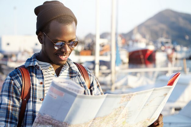 Side view of dark-skinned tourist with backpack in trendy hat and sunglasses examining directions using his paper road map. yacht park or club in picturesque resort town