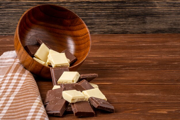 Side view of dark and white chocolate pieces scattered from a wooden bowl on rustic background with copy space