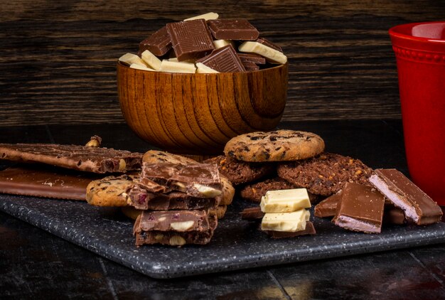 Вид сбоку темного и белого шоколада в деревянной миске и овсяное печенье разбросаны по темному фону