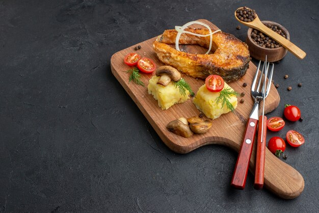 Вид сбоку вкусной жареной рыбы и грибов, помидоров, зелени на деревянной разделочной доске, набор столовых приборов, перец на черной поверхности