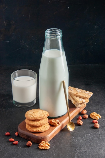 Side view of delicious milk in bottle and glass golden spoon stacked cookies on wooden tray on black background