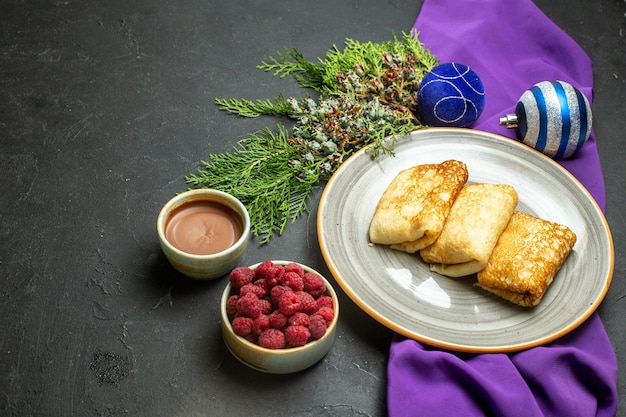 Вид сбоку на аксессуары для украшения вкусных блинов на фиолетовом полотенце и шоколадной малине на черном фоне