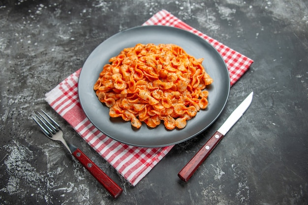 Free photo side view of delicious pasta meal on a black plate for dinner on a red stripped towel and cutlery set on dark background