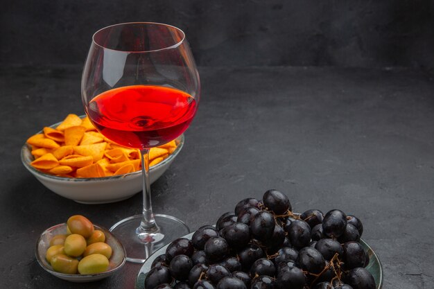 Side view of delicious red wine in a glass goblet and various snacks on a black background