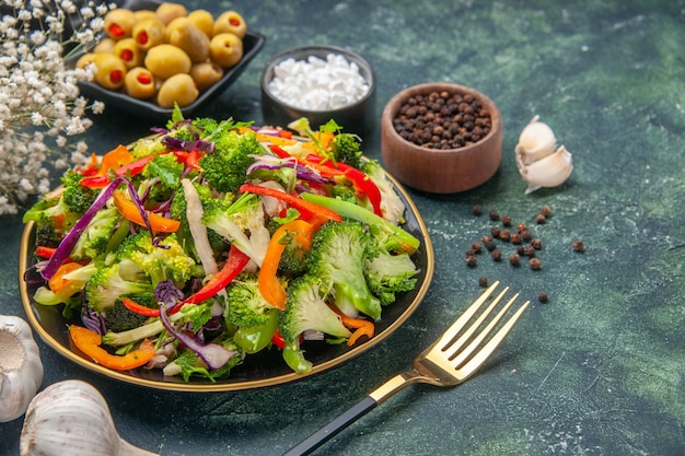 Side view of delicious vegan salad in a plate with various vegetables and fork pepper green black olives garlic on dark background