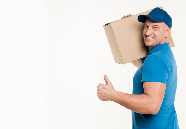 Side view of delivery man carrying cardboard box and giving thumbs up