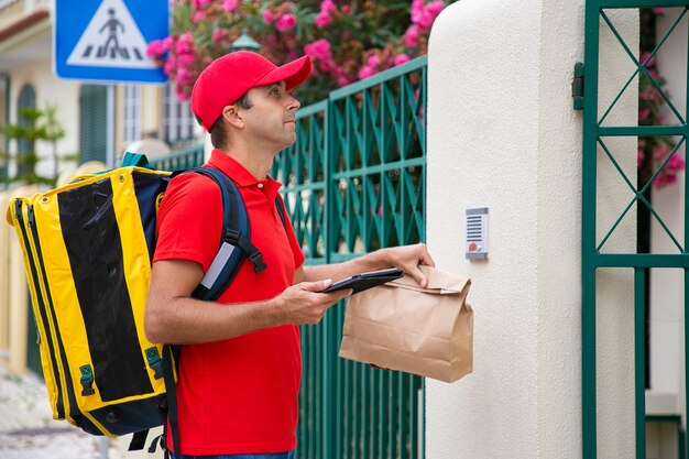 Side view of deliveryman in red cap waiting for customer at entrance. Serious courier with yellow thermal backpack and packet delivering express order to client. Delivery service and post concept