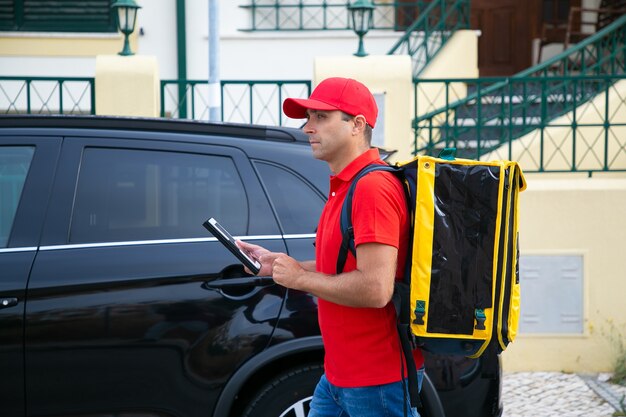Вид сбоку доставщик, наблюдая адрес на планшете. Контент-курьер в красной кепке и рубашке с желтым термосумком доставляет экспресс-заказ пешком. Служба доставки еды и концепция онлайн-покупок