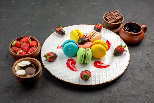 Side view dessert bowls of chocolate strawberries next to the plate of appetizing French macaroons and strawberries on the table