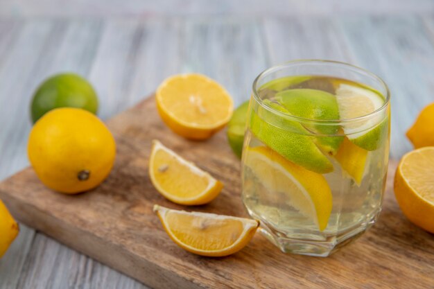 Детокс-вода, вид сбоку в стакане с дольками лайма и половинкой апельсина и лимона на разделочной доске