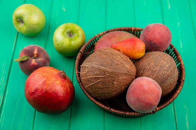 Side view of different fruits such as coconut peach apple pomegranate pear on a bucket on green surface