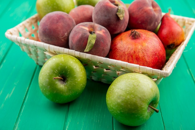 Side view of different fruits such as peach pomegranate on a bucket with green apples on green surface