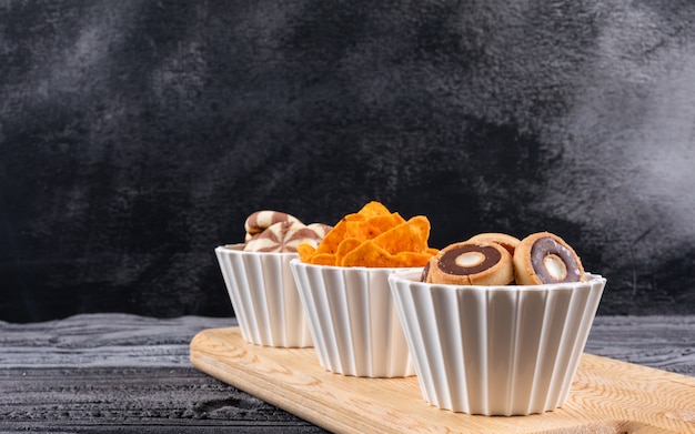 Side view of different kind of snacks as cookies and chips in bowls on cutting board on dark surface horizontal