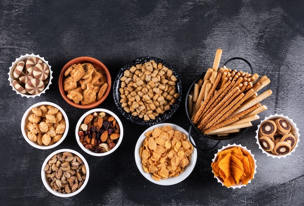 Side view of different kind of snacks as nuts, crackers and cookies in bowls with copy space on dark surface horizontal