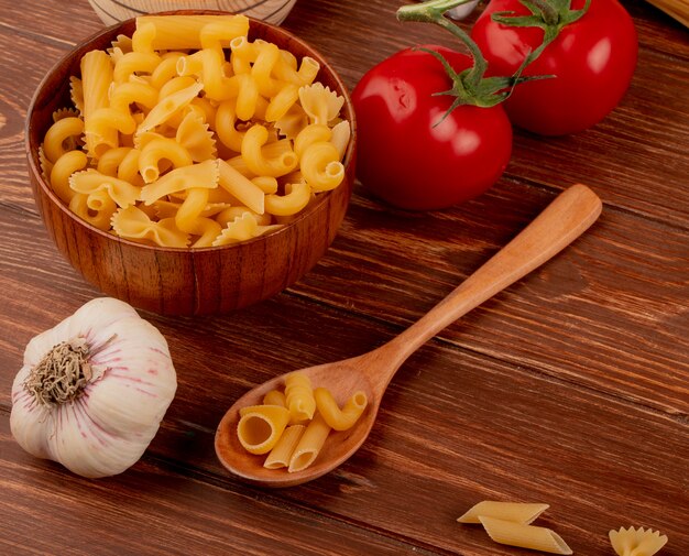Side view of different macaronis in wooden bowl and spoon with garlic and tomatoes on wooden table