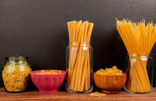 Side view of different types of macaroni in jars and bowls on wooden surface and black surface