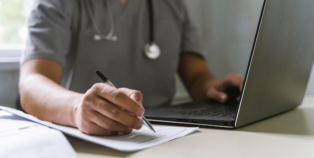 Вид сбоку на врача со стетоскопом, работающего на ноутбуке и пишущего на бумаге